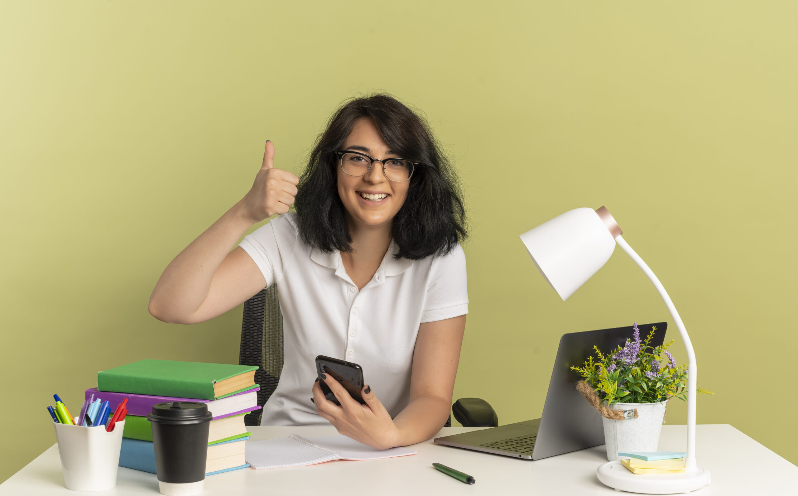 young smiling pretty caucasian schoolgirl wearing glasses sits at desk with school tools thumbs up and holds phone isolated on green background with copy space